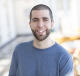 White male, around age 30, with brown buzz cut hair and a dark brown beard. He has light blue eyes, and is standing outside smiling and wearing a light blue crew neck sweater. 