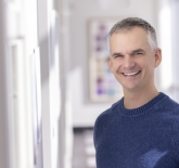 middle aged, white male, with buzz cut white and grey hair. He is wearing a navy blue crew neck knit sweater, and it standing in a hallway smiling.