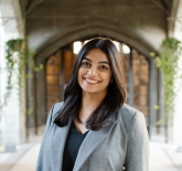 a woman, with brown skin tone, with shoulder-length dark brown hair. She is wearing a slate grey pea-coat, and a black v-neck undershirt. She is standing and smiling in front of a cement archway. 