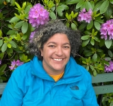 woman with dark and light grey curly hair, light brown skin, and is wearing a bright blue windbreaker with a yellow t-shirt underneath. There are pink/purple flowers and greenery in the back. 