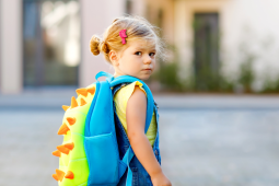 A young child with blonde hair in a bun, wearing denim overalls and a yellow shirt, looks over their shoulder while carrying a bright blue and green dinosaur backpack with orange spikes.