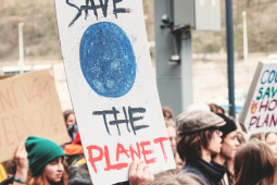 A group of people holding signs at a rally. One sign says "SAVE THE PLANET" with a blue drawing of the Earth. Another sign says "COOL KIDS SAVING HOT PLANET".
