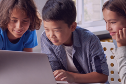The image shows three children looking at a laptop computer with an intrigued, concentrated expression.