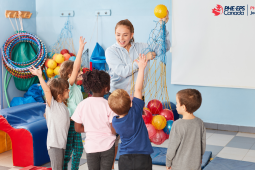 A teacher holding a net with balls in it in a room that seems to allow physical activity for pupils. She has 5 pupils in front of her, 3 girls and 2 boys. 3 of them raise their hands to get a ball from the teacher. / Une enseignante qui tient un filet avec des ballons dedans dans une salle qui semble permettre de l’activité physique à des élèves. Elle a en face d’elle 5 élèves, 3 filles et 2 garçons. 3 d’entres eux lèvent la main pour avoir une balle de la part de l’enseignante.