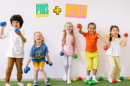 A group of kids are standing against a white wall, smiling and holding colorful balls ready to throw.