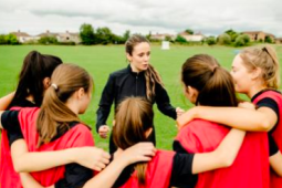 group of 5 young women in a huddle wearing red jerseys and a coach in the centre wearing a black jacket on a soccer field