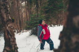 man standing on snow in the woods looking up wearing a red winter jacket. 
