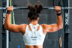a woman holding up a barbell above her head with weight plates on the ends. She is wearing a white sports bra and has her brown hair up in a bun. 