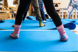 a young student standing on a blue yoga mat in their classroom. You see from their knee down. They are wearing black tights and pink and blue socks. The yoga mat is in the classroom with other students doing the same. 