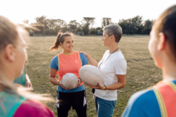 a women coach/teacher talking to adolescent girls. They are holding rugby balls and wearing pinnies while standing in a grass field outside. 