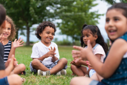 5 young children sitting cross legged in a grass field counting on their fingers