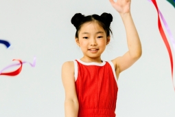 A young person in a red leotard smiles while holding up a hand and waving colorful ribbons against a white background.