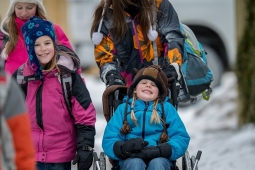 child wearing a blue winter jacket in a wheelchair being pushed by an adult. to young children on the left with a pink and orange jacket. all in the snow. 