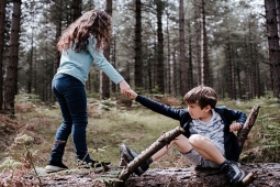 two children in the forest. one is on the ground and the other is helping them up by holding their hand. 