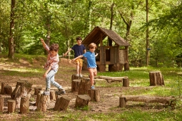 3 children walking on tree stumps in a forest. There is a tree fort behind them.