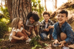 4 children sitting down in the forest. There are two little girls and two little boys. The boys are looking forward smiling and the two little girls are smiling at each other. 
