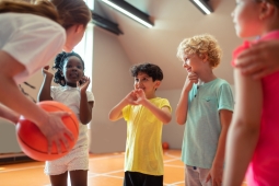 a teacher leaned over talking to 4 of her students during gym class. She is holding a orange basketball and they are all standing in a gymnasium.