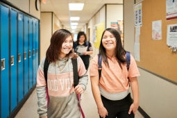 two young indigenous girls walking down a hallway in a school. They both have a backpack on and are smiling. There is a set of blue lockers next to them and a cork display board on the other wall. 