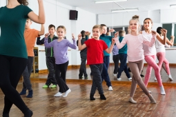 many young students dancing in a dance studio. There is an instruction in front of them wearing a green shirt and black pants. There is a mirror behind the students. 