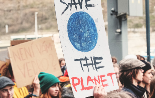 A group of people holding signs at a rally. One sign says "SAVE THE PLANET" with a blue drawing of the Earth. Another sign says "COOL KIDS SAVING HOT PLANET".