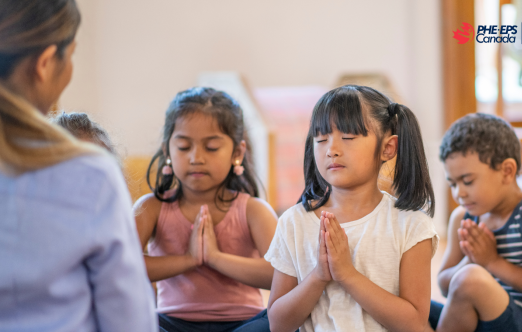 The image shows three children practicing mindfulness with praying hands, eyes closed, in front of their teachers.