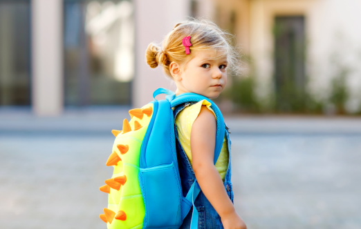 A young child with blonde hair in a bun, wearing denim overalls and a yellow shirt, looks over their shoulder while carrying a bright blue and green dinosaur backpack with orange spikes.