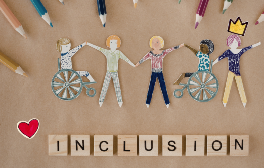Paper cutouts of diverse people, including some in wheelchairs, holding hands, surrounded by colored pencils. Wooden letter blocks spell out 'INCLUSION'.
