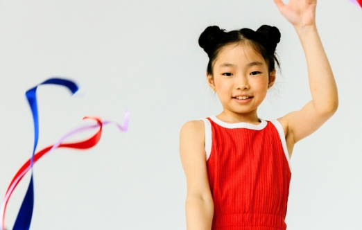 A young person in a red leotard smiles while holding up a hand and waving colorful ribbons against a white background.