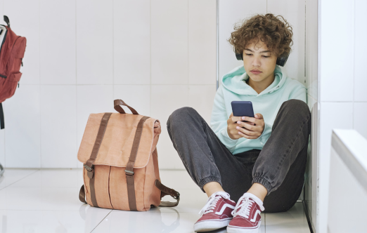 A teenage boy is sitting on the floor with a schoolbag next to him. He is holding a phone and looking at it intently.